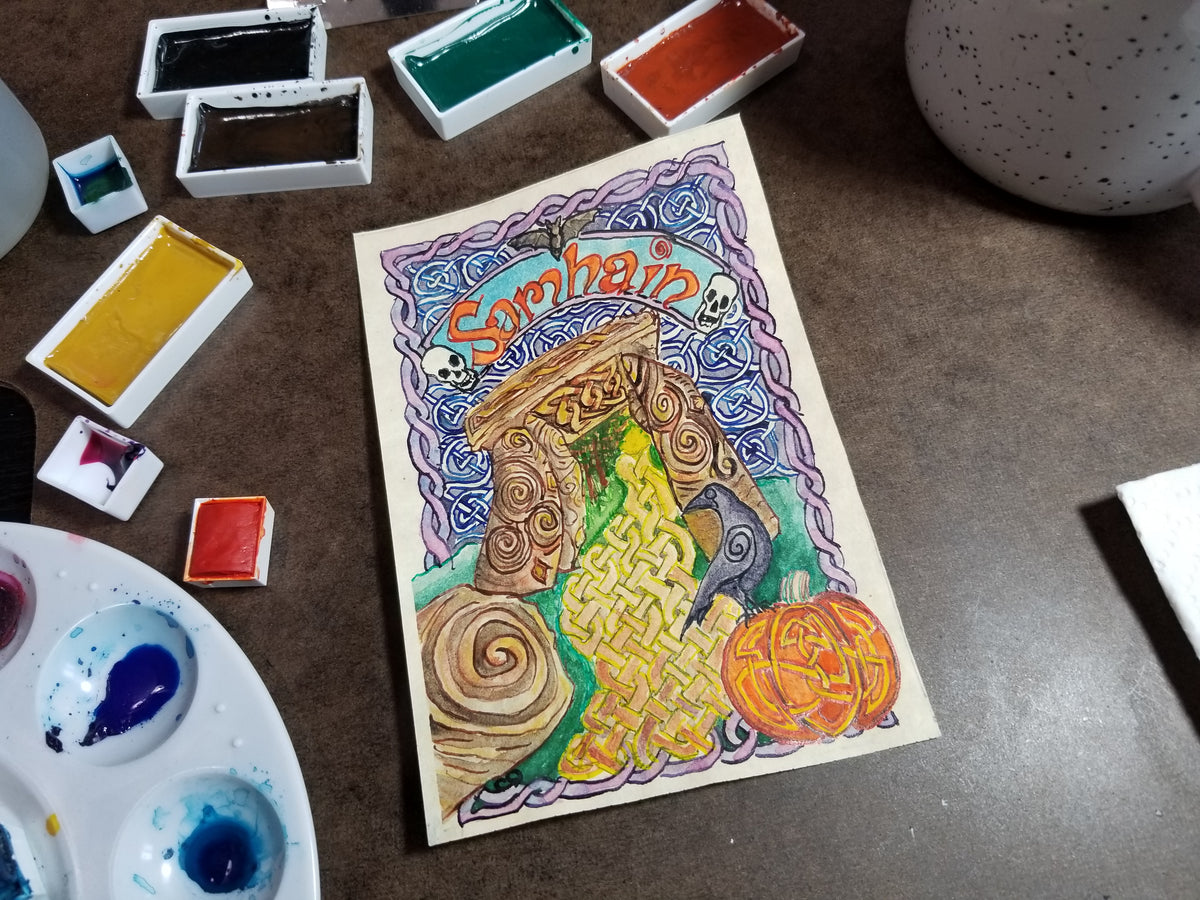 My Samhain mini painting featuring carved standing stones, a pumpkin, a crow, and a misty landscape, displayed on a brown tabletop surrounded by paint palettes and paint tubes.
