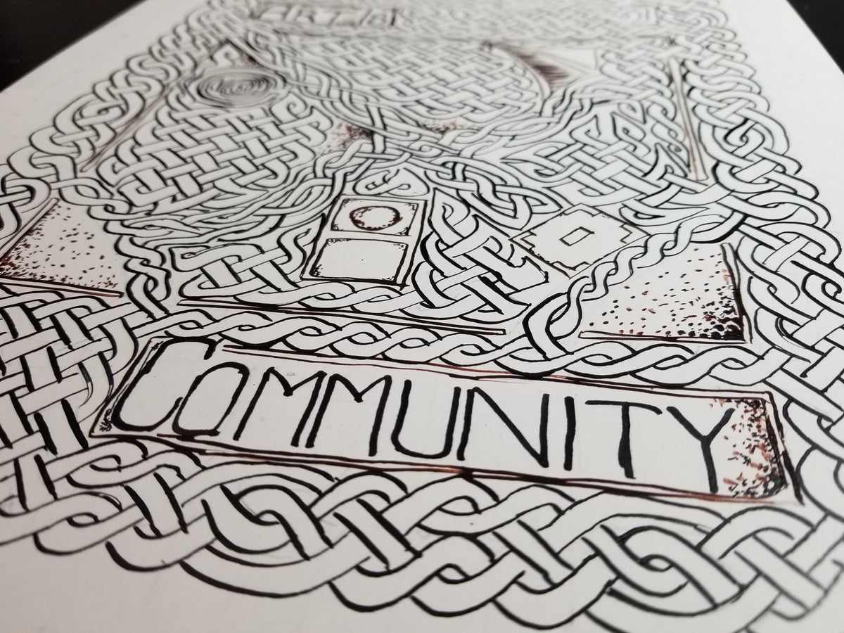 A close-up of the inking stage of the painting Art is Community, a black ink illustration on white paper, featuring intricate Celtic knot patterns and the word 'COMMUNITY' in a banner across the center.