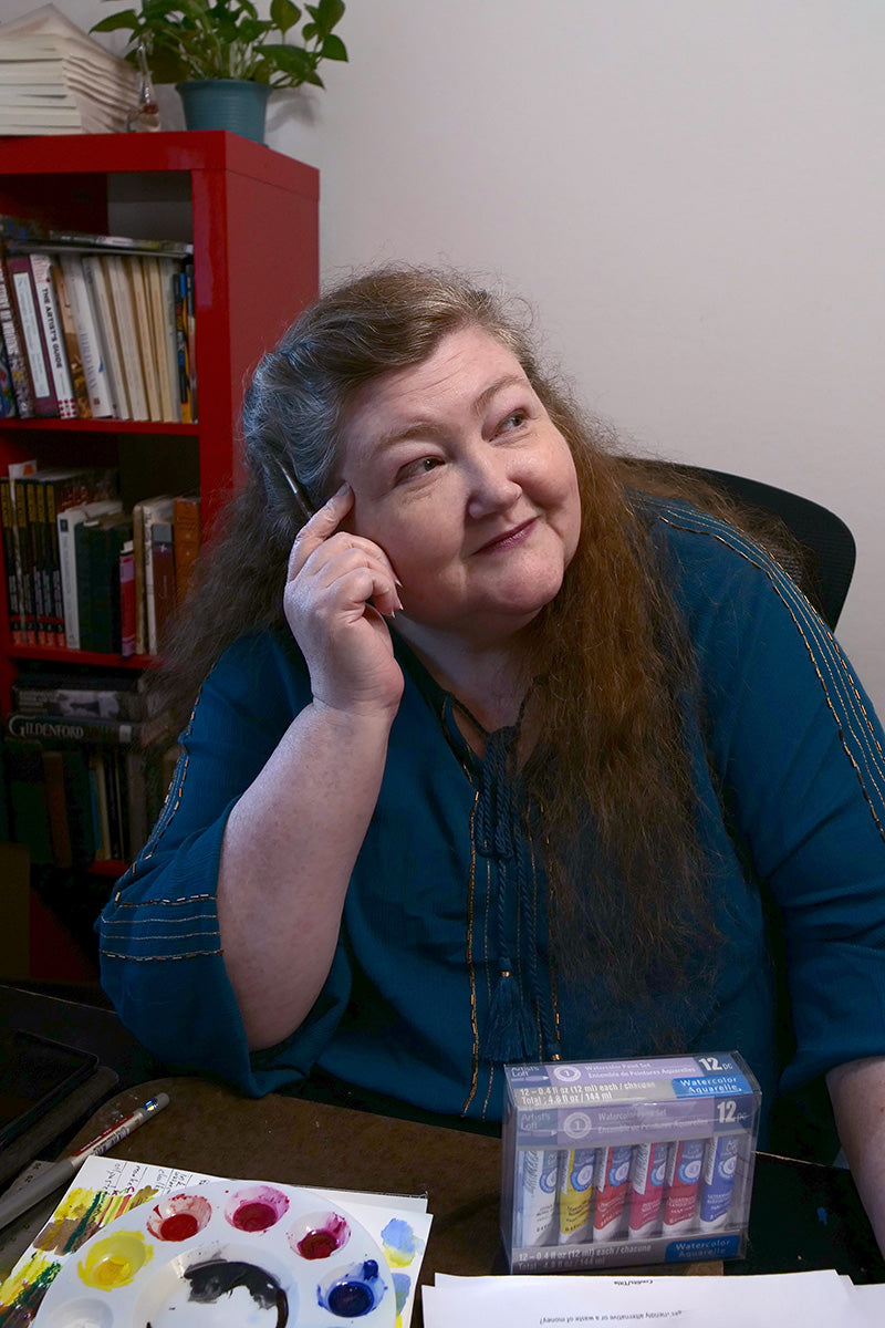 Crys Odenkirk, a woman with long red hair, sits at a desk wearing a blue beaded shirt, leaning her head on one hand, which is holding a paintbrush. In front of her is a paint palette with mixed colors and a box of watercolor paints.