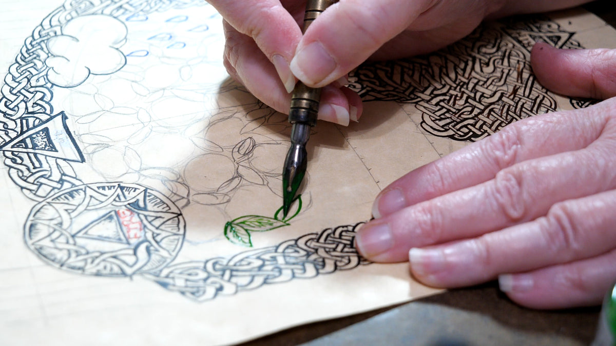 Close-up of a person's hands using a green ink calligraphy pen to draw green leaves on a piece of beige paper with a black Celtic knot design.